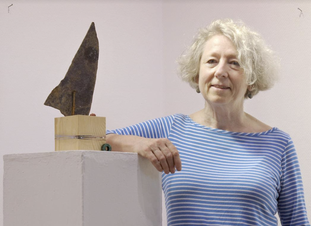 photo of woman with grey hair, striped shirt next to a small sculpture