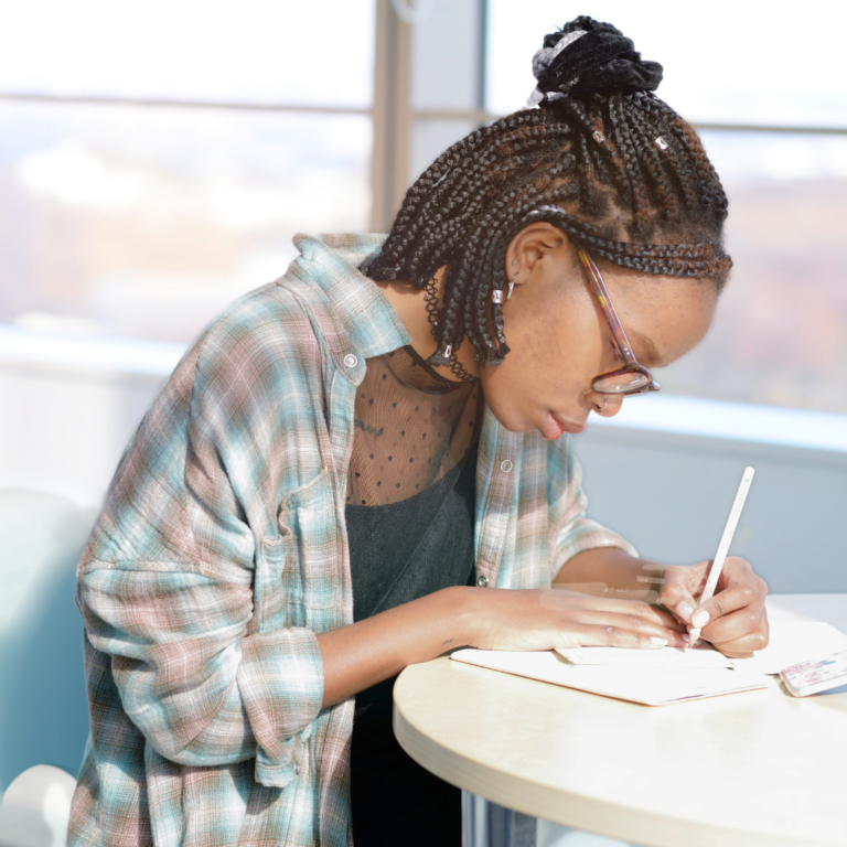 Artist Meetups image of Young black woman writing in a notebook with a neutral office window background.