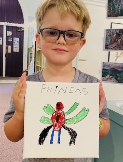 youth approx ages 6-7 with glasses blond hair, holding artwork they created of a colorful, cute insect.