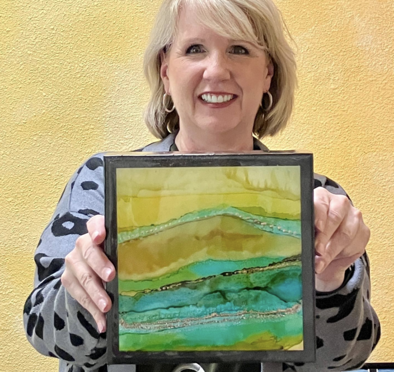 artist with blonde hair holding an 8x8 artwork with abstract greet and gold colors